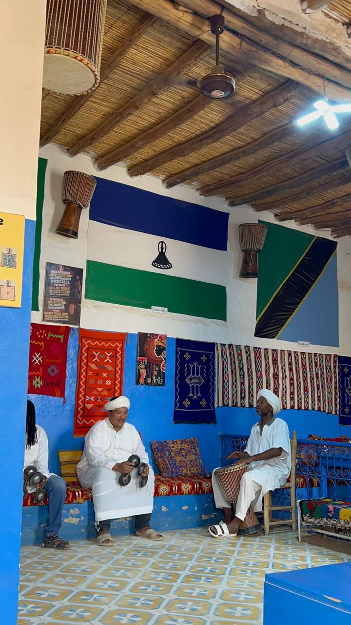 Interior scene with colourful textiles and flags, and two men in traditional attire seated below.