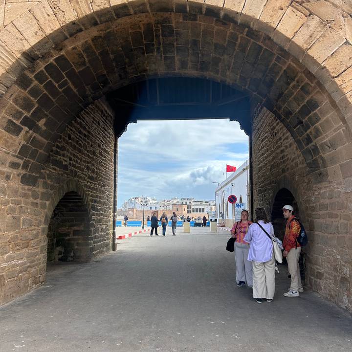 Visitors stand under an old stone archway looking out to a coastal Moroccan town and cloudy sky.