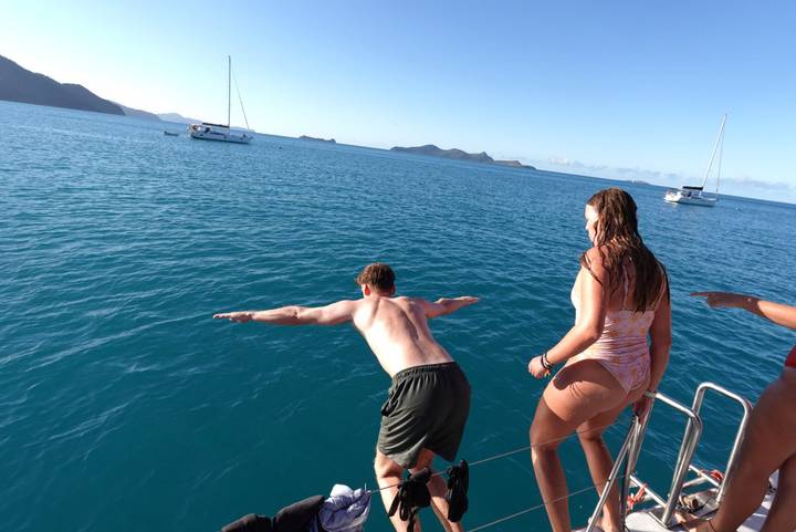 From a sailboat, a man dives into turquoise water while a woman watches on a sunny day.
