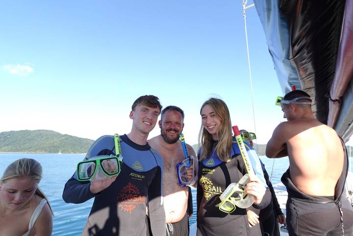 Travellers in wetsuits hold snorkel gear aboard a sailboat with an island backdrop.