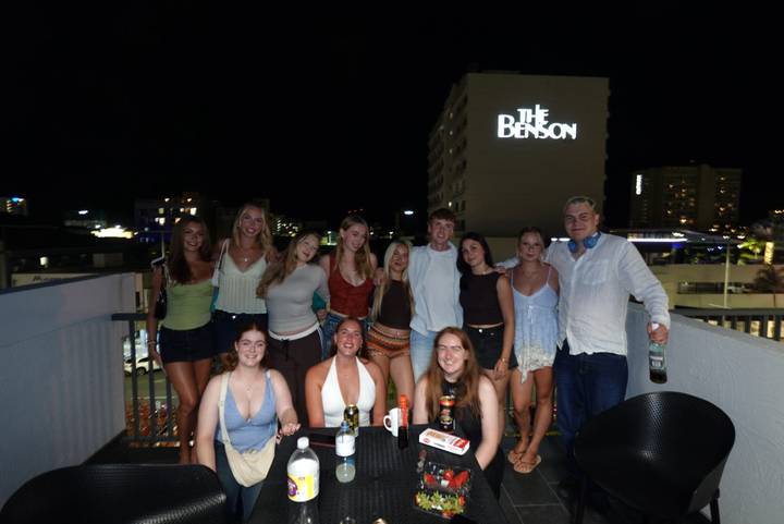 Nighttime rooftop gathering of friends with city lights and a hotel sign reading 'The Benson' in the background.