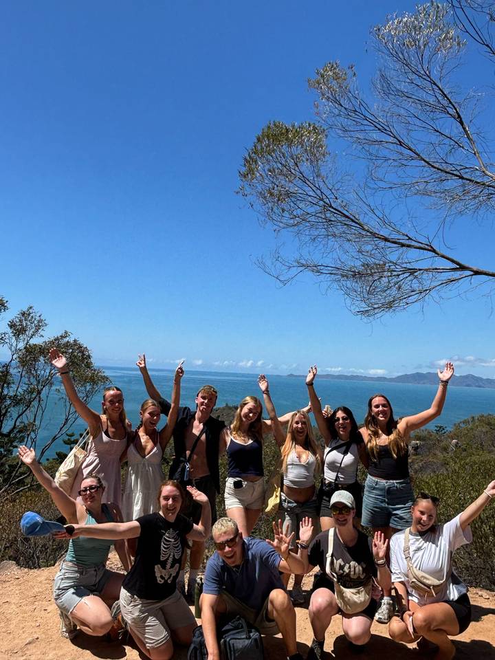 A line of travellers raise their arms in celebration at a coastal lookout with blue sea behind.