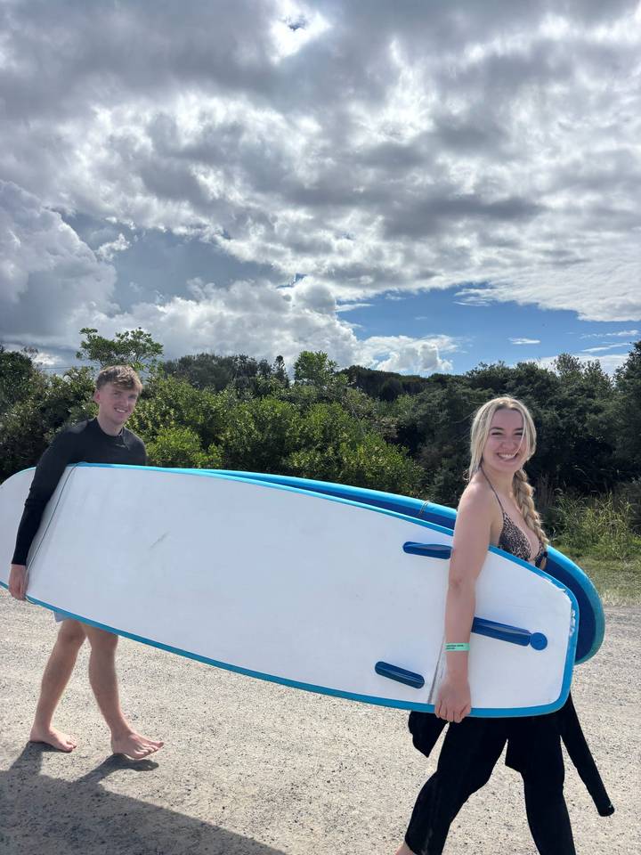 Two surfers carry a large blue-rimmed surfboard through leafy greenery under a bright sky.