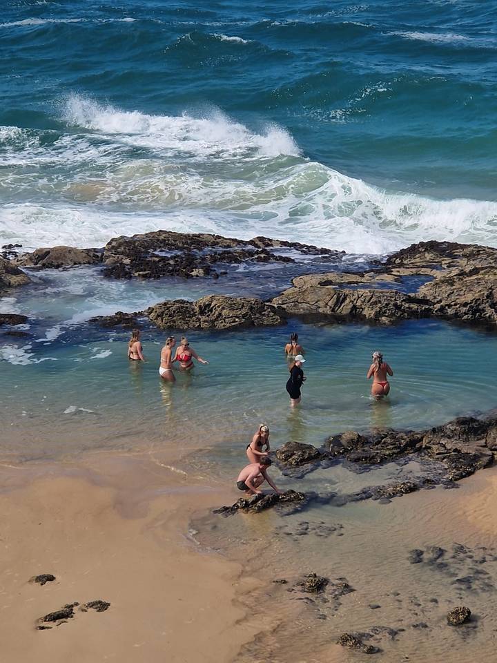 Travellers wade and swim in a clear natural rock pool beside crashing ocean waves.