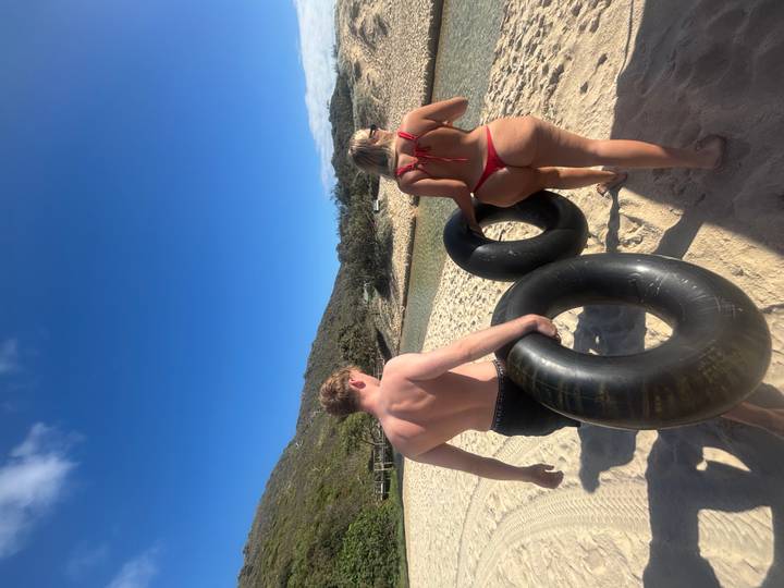 A man and woman carry large inner tubes down a sunny sandy beach toward a creek.