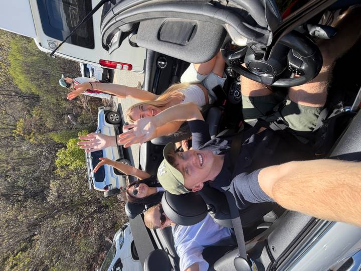Group riding in a convertible cheer with raised arms on a sunny scenic road.