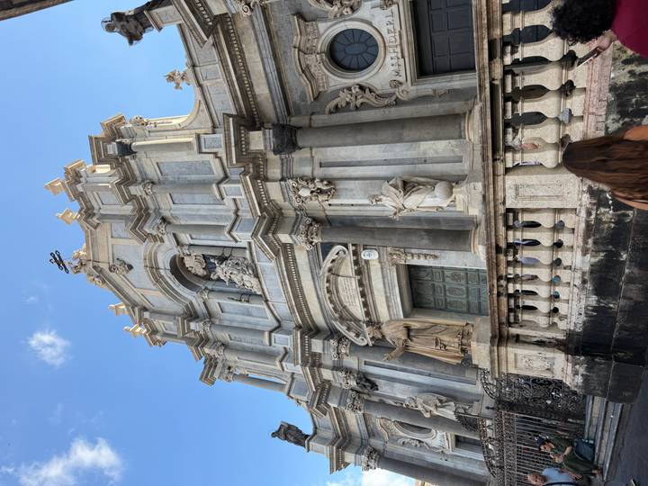 Impressive grey-stone baroque church façade with statues and columns set against a blue sky, visitor walks past.