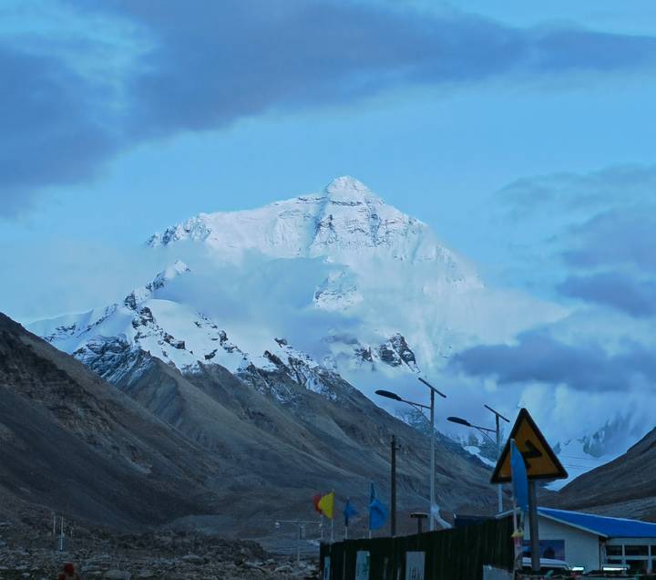 Snow-clad Himalayan peak emerging through clouds above rugged foothills at dusk-blue hour.