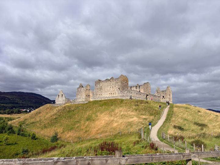 Ruins of a stone castle (Ruthven Barracks) sitting atop a grassy mound under a grey sky with visitors walking up.