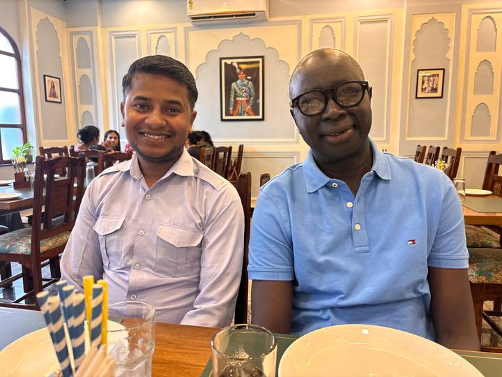 Two men smiling while seated inside a traditional Indian restaurant decorated with Mughal artwork.