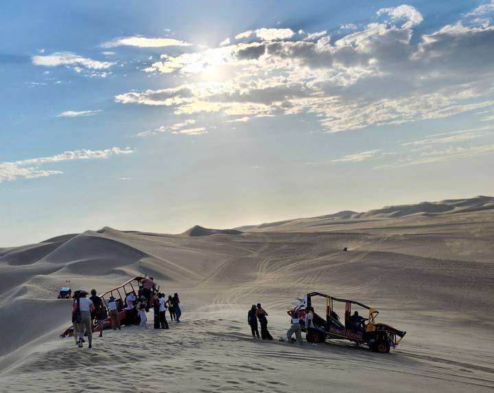 Tourists gather around sand-buggies on vast sun-lit dunes under a hazy afternoon sky.
