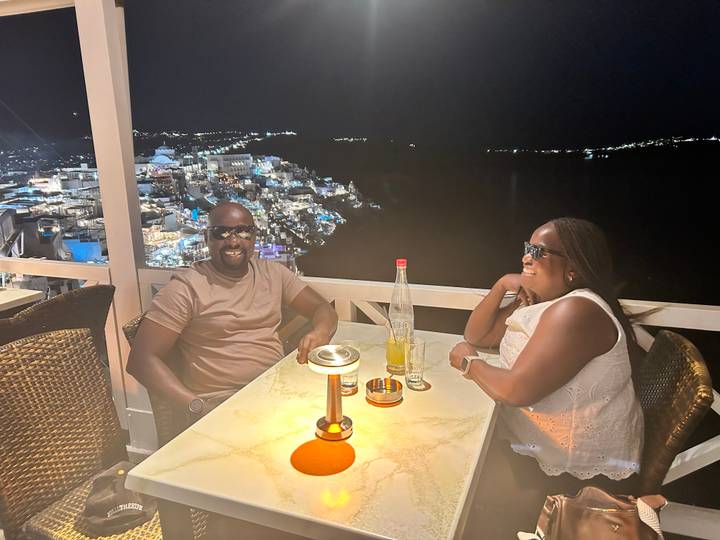 Couple enjoys candle-lit dinner on a balcony overlooking a glittering island town at night.