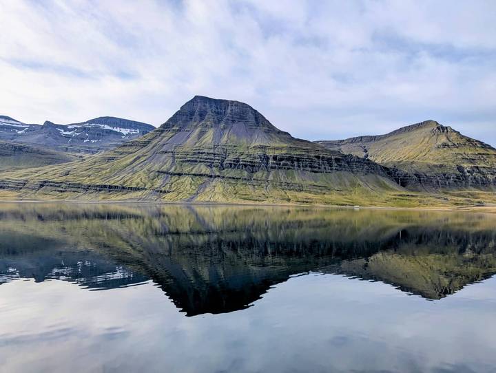 Craggy green mountain reflected perfectly in a calm fjord under a pale sky in Iceland