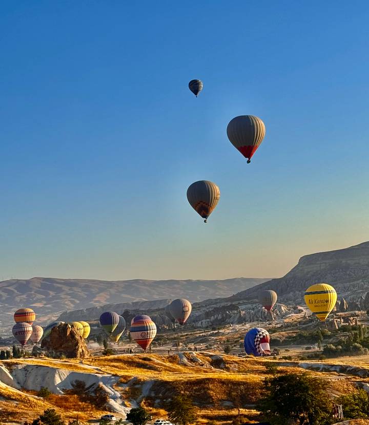 Hot-air balloons rise high above layered hills in the soft early morning blue sky.