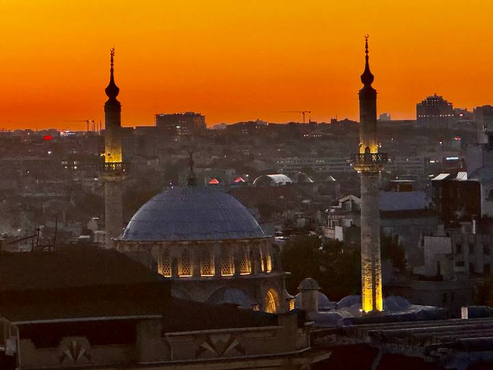 Closer view of illuminated mosque dome and twin minarets against vivid orange twilight sky.