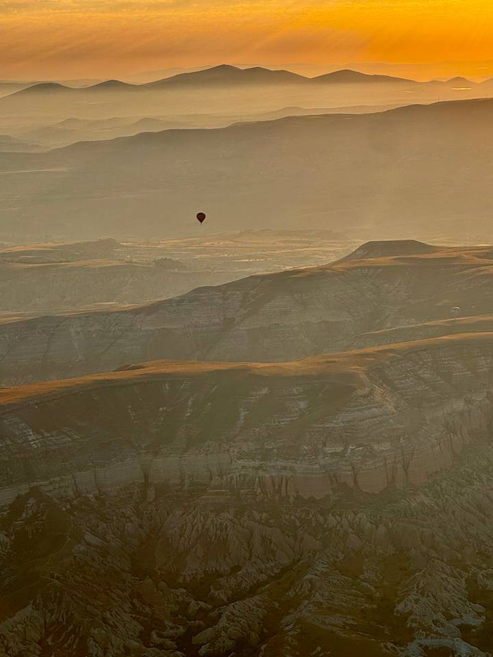 Single hot-air balloon drifts above layered golden valleys in hazy morning light.
