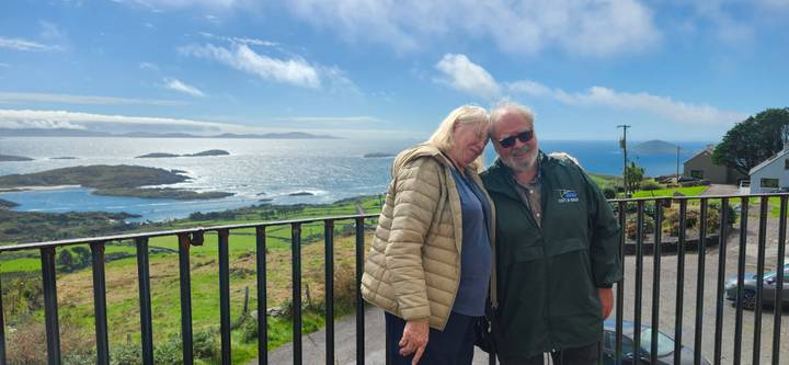 Smiling couple poses on a balcony overlooking sparkling sea, islands and rugged coastline under blue sky.