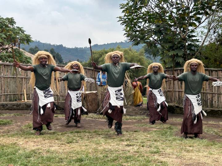 Traditional Rwandan dancers in green shirts and cow-hide skirts leap energetically holding spears in village setting.