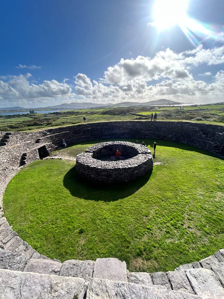Circular stone ring-fort structure with tourists exploring inside and atop the walls amid coastal Irish scenery