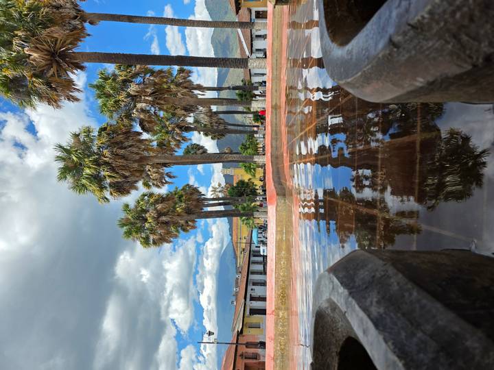 Tall palm trees line a reflective fountain pool in a colonial plaza backed by rolling hills and dramatic clouds.