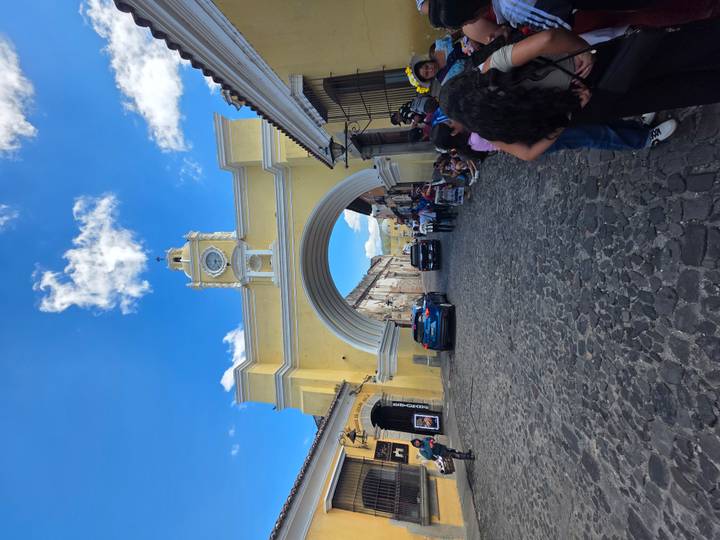 Tourists pass beneath the iconic yellow Santa Catalina Arch in Antigua with a volcano peeking through the archway under a vivid blue sky.