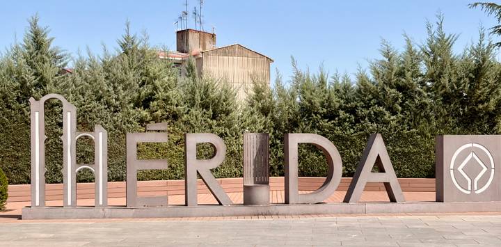 Large modern sculpture spelling MERIDA set before tall shrubs under a clear sky.