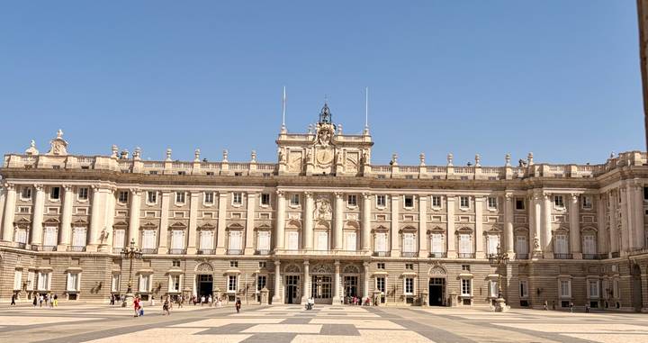 Wide façade of an ornate royal palace overlooking a spacious courtyard with scattered visitors.