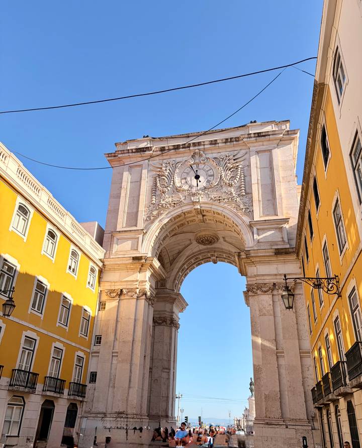 Elegant arch with clock and baroque carvings framed by bright yellow buildings under blue sky.