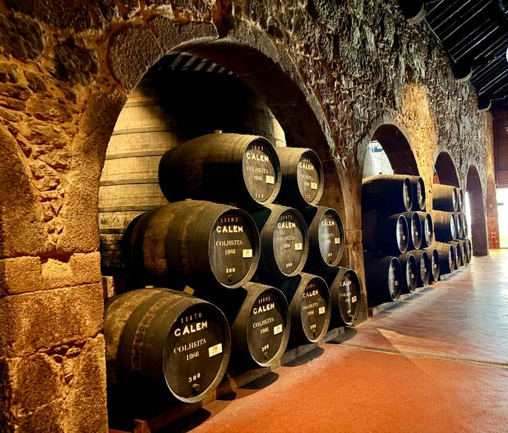 Rows of stacked wooden port barrels inside a dimly lit stone cellar.