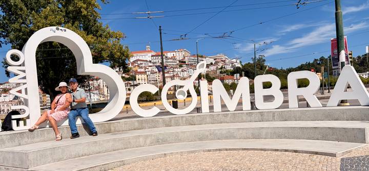 Couple posing beside large white COIMBRA sign with hilltop cityscape in background.