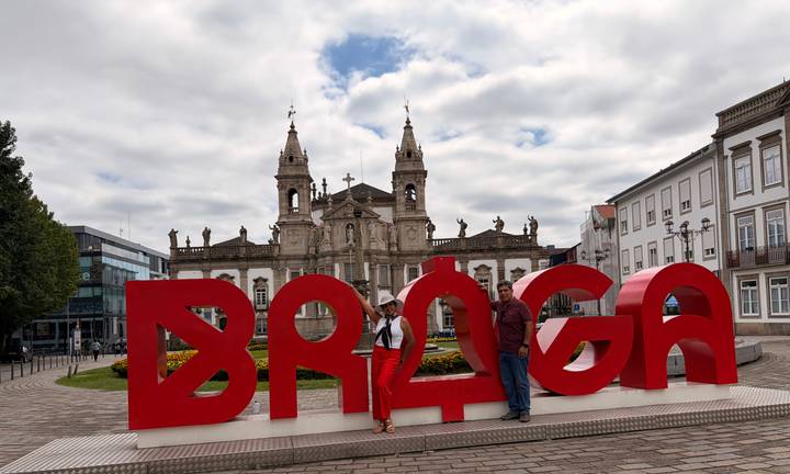 Two travelers stand by bold red BRAGA letters with historic church behind.