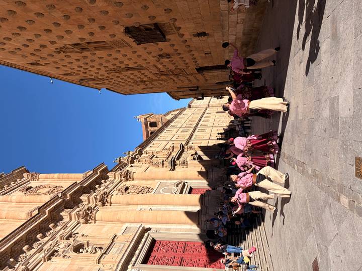 Traditional dancers in pink costumes perform in a stone plaza between historic buildings.