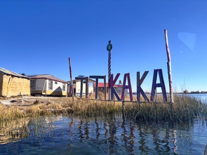 Large colourful letters spelling "TITIKAKA" standing in reeds along the calm lakeshore under clear blue sky.