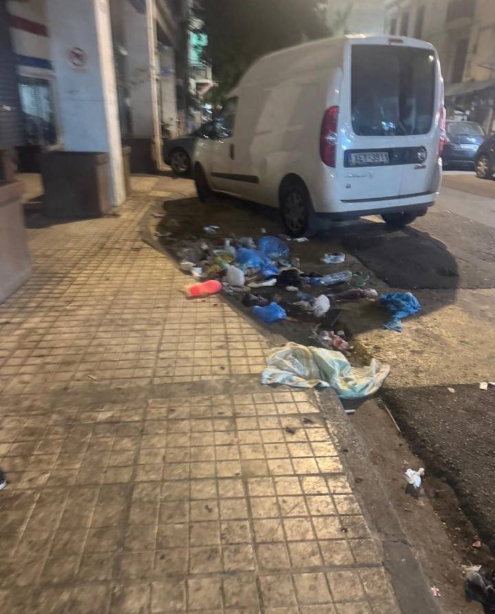 Nighttime city sidewalk scattered with rubbish beside a parked car.