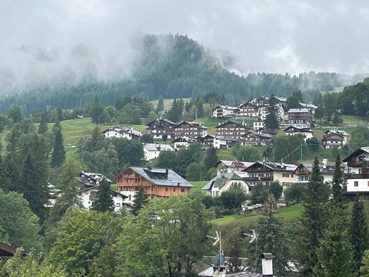 Misty alpine village with wooden chalets scattered across rolling green hills and conifer forests.