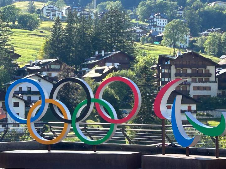 Colorful Olympic rings sculpture set against alpine village houses and green hills in bright sunlight.