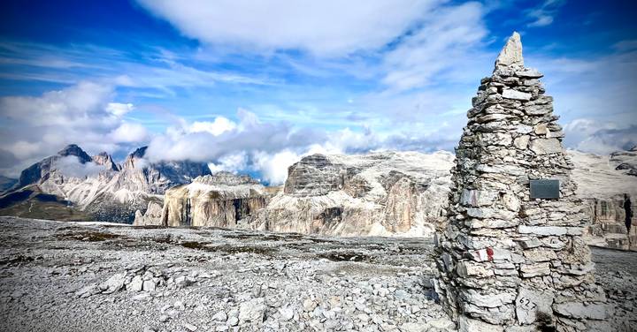 Rock-strewn high-altitude plateau in the Dolomites with a tall stone cairn foreground and dramatic rocky peaks beyond.