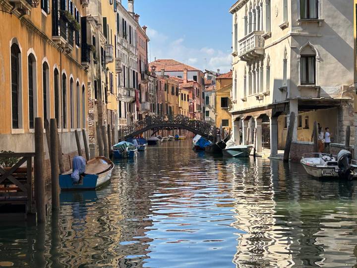 Quiet Venetian canal lined with colorful buildings and small boats, crossed by an arched pedestrian bridge.