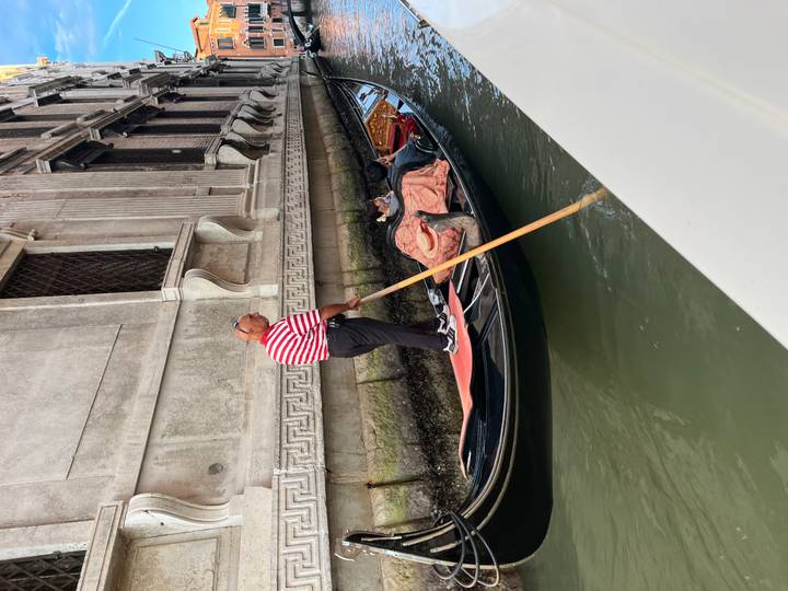 Striped-shirt gondolier steering a gondola with passengers through a narrow Venetian canal beside historic palaces.
