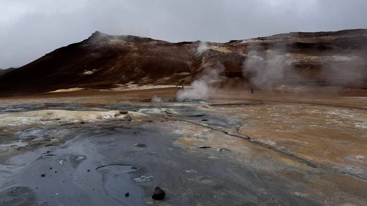 Barren geothermal field with steaming fumaroles and mineral-stained earth beneath gray skies.