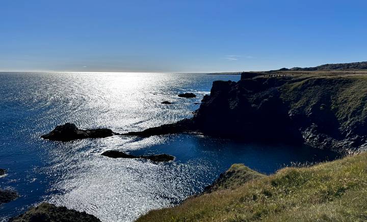 Sun-lit Atlantic coastline with steep grassy cliffs and sparkling blue sea near Arnarstapi.