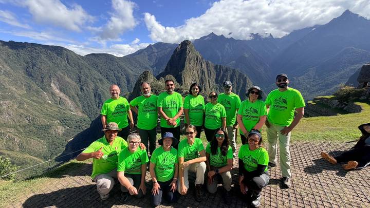 Large trekking group in matching green shirts posing with Machu Picchu citadel and Andean peaks behind.