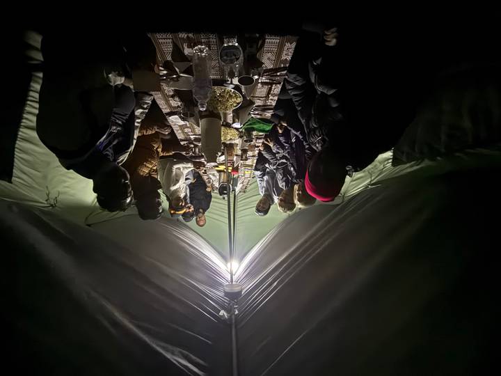 Night-time meal inside a camping tent, trekkers gathered around a long table lit by a central lamp.