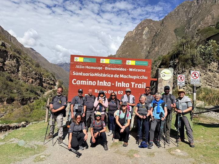 Full trekking group with poles posing at Inca Trail entrance sign in bright sunshine.