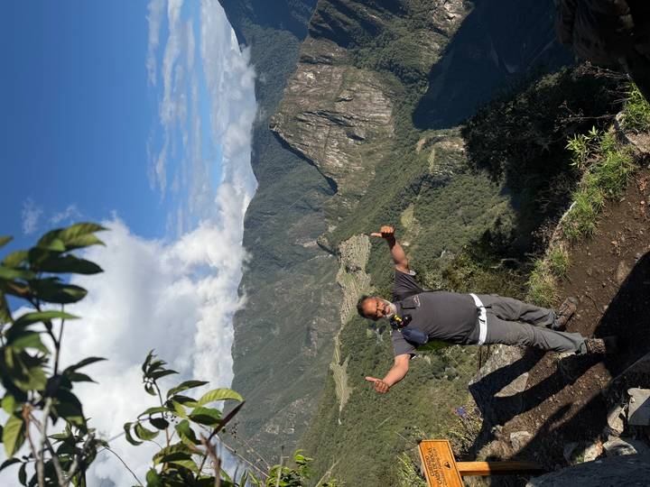 Solo trekker giving thumbs up on a cliff viewpoint overlooking Machu Picchu ruins and green valleys.