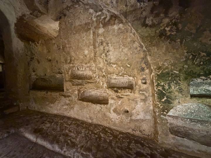 Rough-textured stone wall with several carved tomb niches inside an ancient underground catacomb.