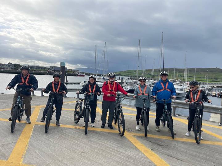 Cycling group lined up with bikes on a harbour pier with sailboats and hills behind.