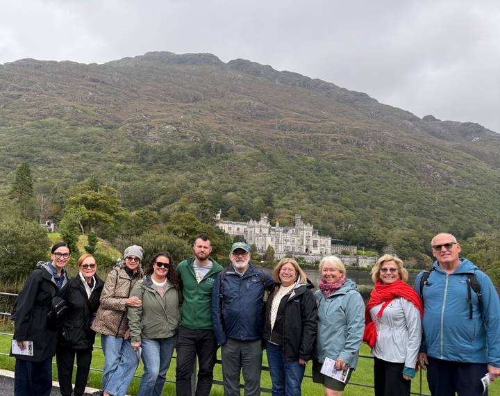 Group of travelers posing in front of a lakeside neo-gothic abbey surrounded by green mountains under a cloudy sky
