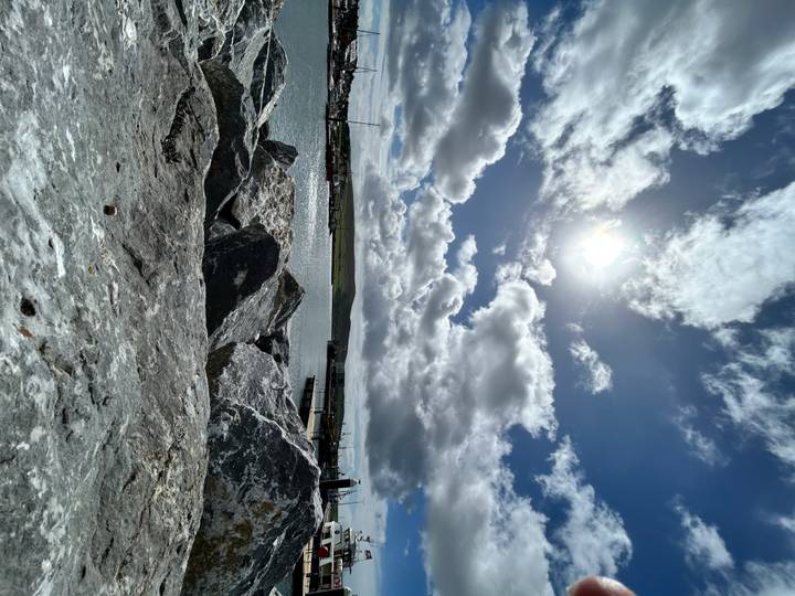 Rocky harbor foreground with bright sun shining through dramatic clouds over calm water and distant pier