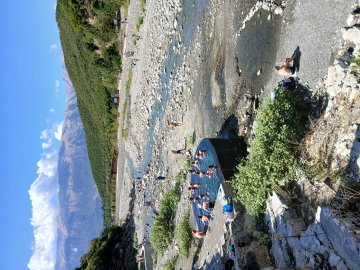 Natural hot springs pools beside a rocky river valley with bathers and mountains in background.